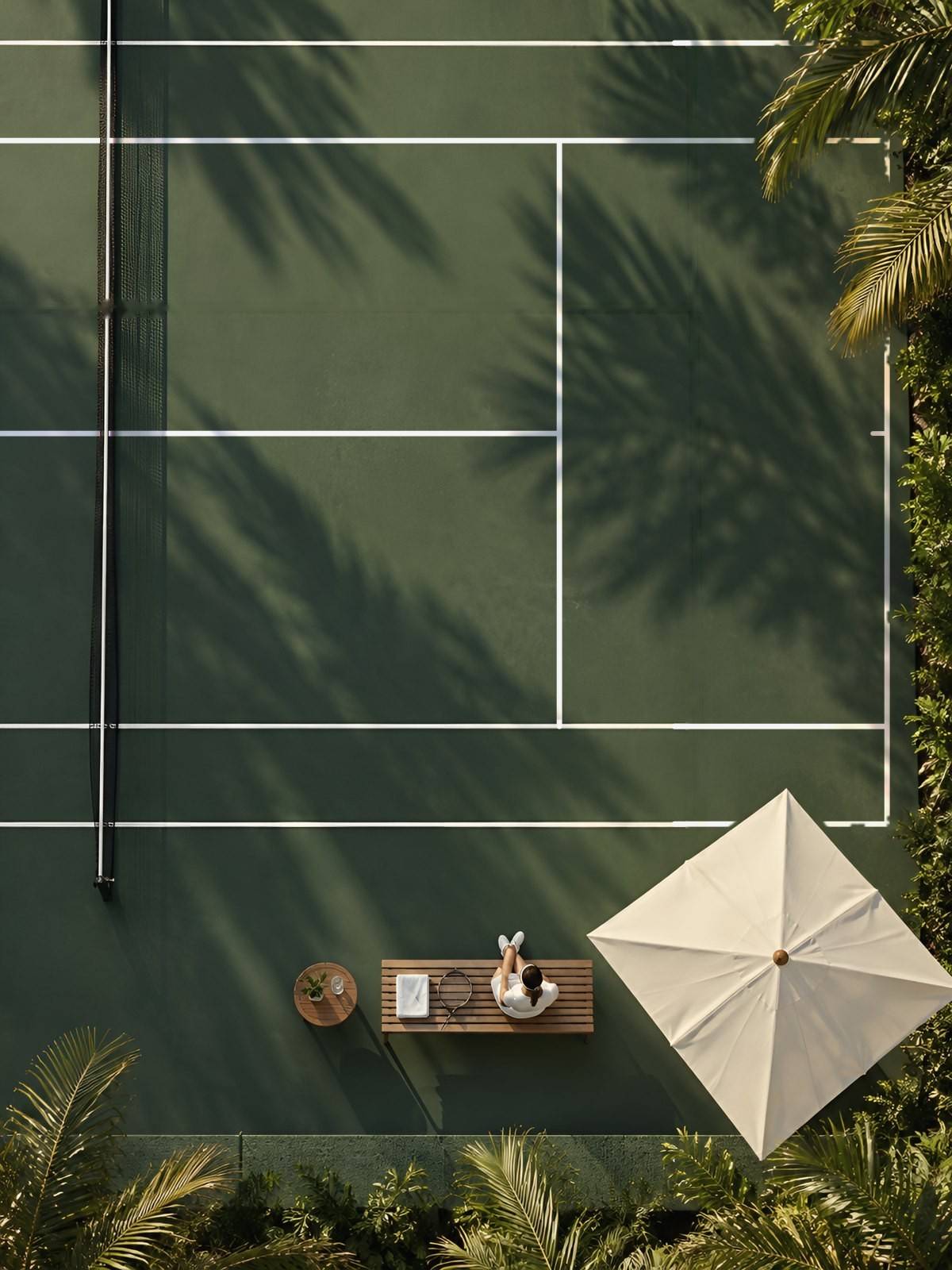 Overhead view of a green tennis court with palm shadows, a parasol and a player resting on a teak bench