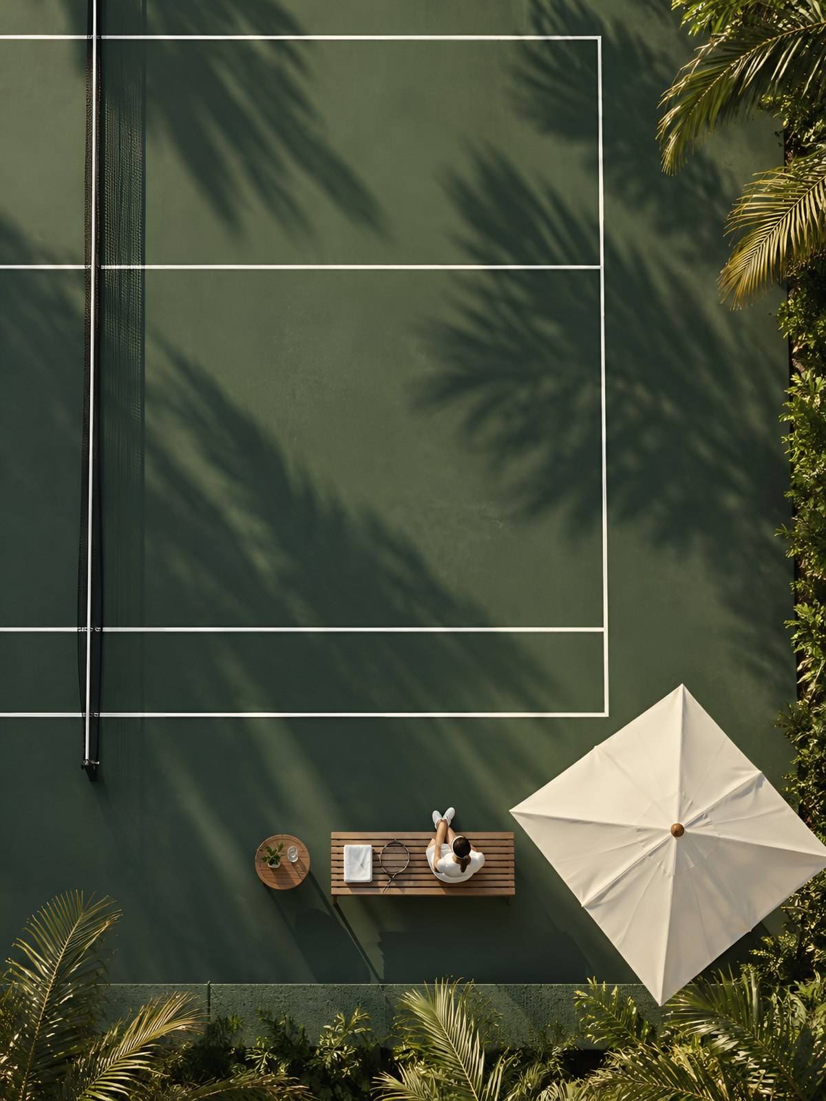 Overhead view of a green tennis court with palm shadows, a parasol and a player resting on a teak bench